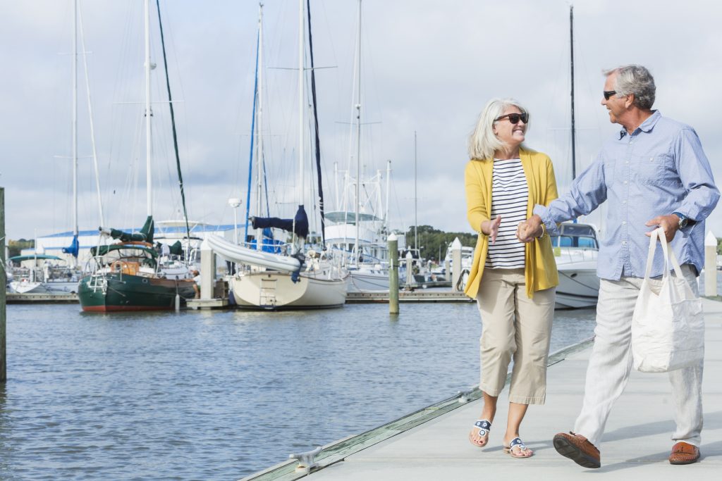 A happy, senior couple walking together holding hands along a boat dock, enjoying the water and view of the luxury sailboats on a sunny summer day. They are smiling, face to face, enjoying retirement.